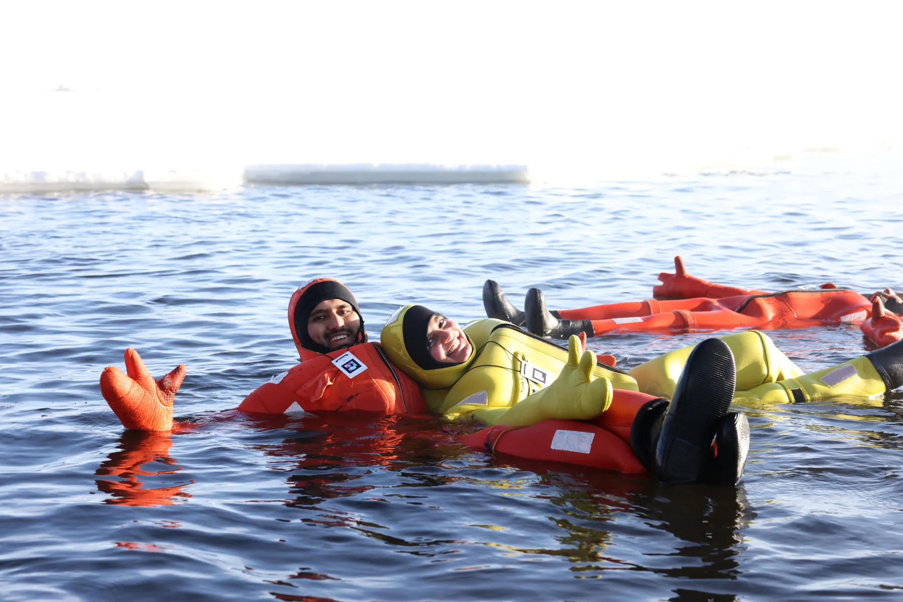 Two people smiling and floating in cold water wearing survival suits, one orange and one yellow.