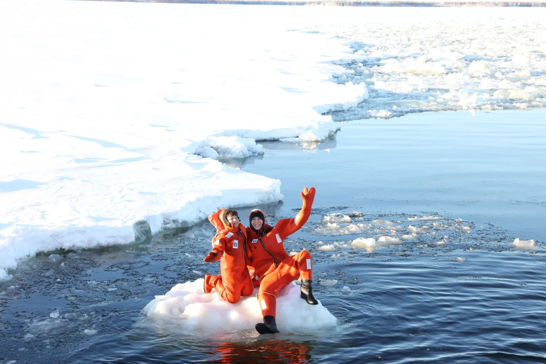 Two people in orange survival suits sitting and waving on a small ice floe surrounded by icy water.