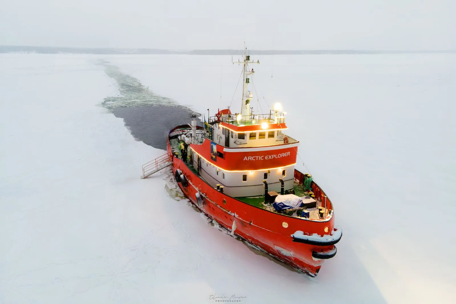 Red and white Arctic Explorer icebreaker ship cutting through frozen icy sea with a clear path behind it during winter.