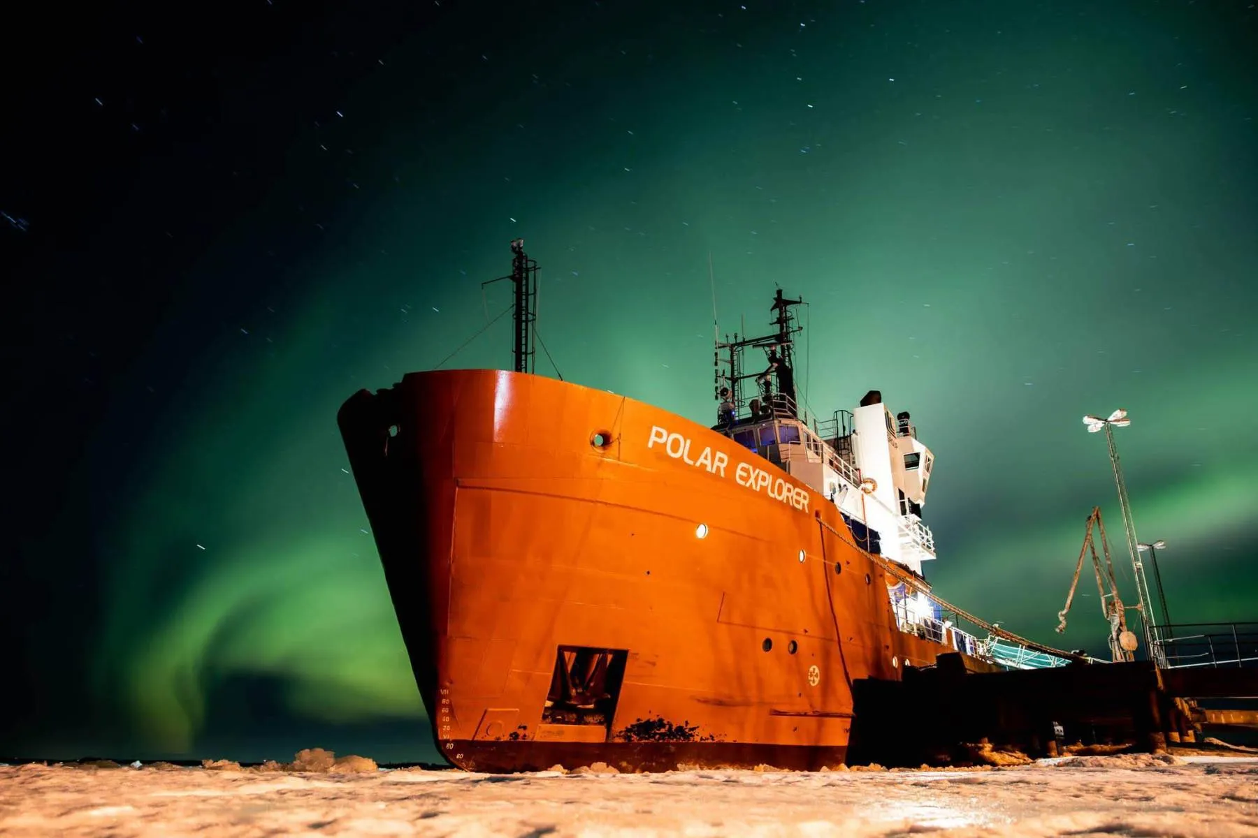 The bright orange Polar Explorer ship docked on icy ground under a vibrant green northern lights sky at night.