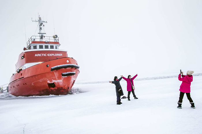 Red icebreaker ship named Arctic Explorer on ice with three people, two posing and one taking a photo.