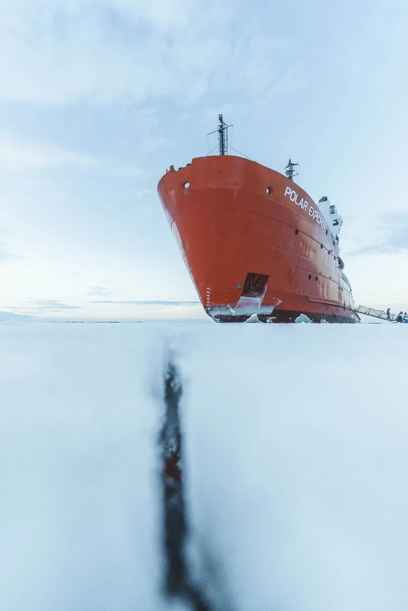 Red icebreaker ship named Polar Explorer stationary on frozen sea with a crack in the ice in the foreground.