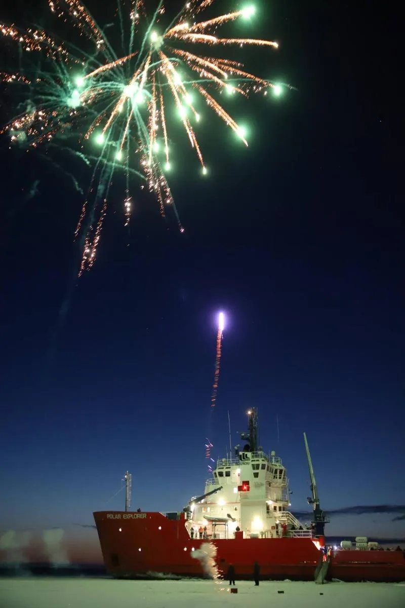 Red icebreaker ship named Polar Explorer on ice with fireworks exploding in the dark night sky above.