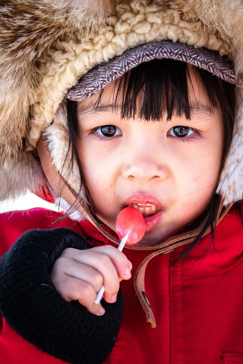 Close-up of a child wearing a fur-lined hat and red coat licking a red lollipop.