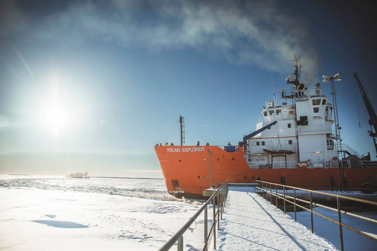 Orange ship named Polar Explorer docked at a snow-covered pier under a bright sun in a clear blue sky.