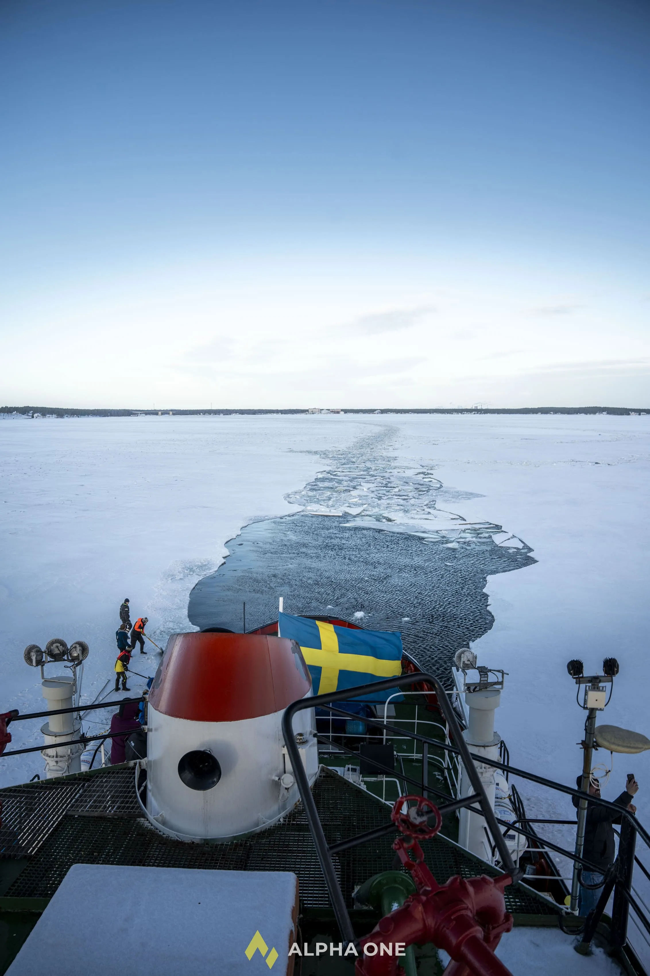 View from an icebreaker ship's deck breaking through a frozen sea with a Swedish flag, while crew members work on the ice.