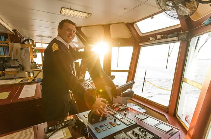 Ship captain operating controls in the bridge with sunlight streaming through the windows.
