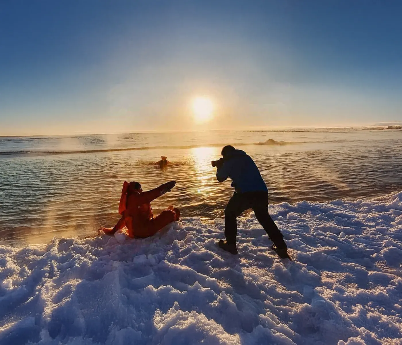 Photographer in dark clothes taking pictures of a person in an orange suit sitting on snowy ground near a steam rising from icy water at sunset.